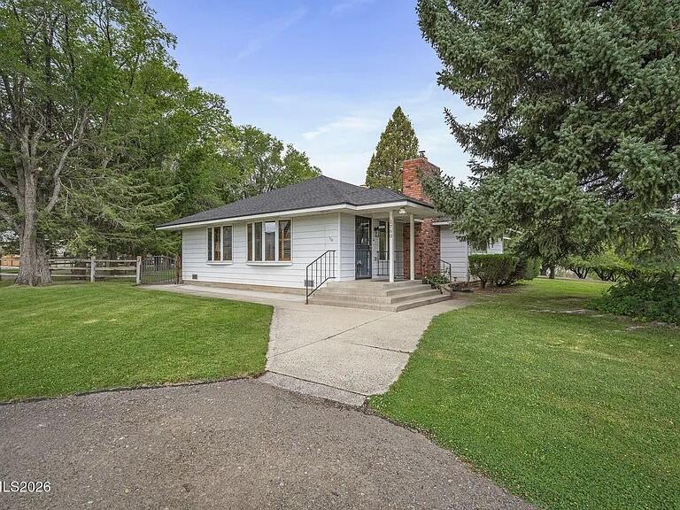Front of the 1950s farmhouse, white siding with a brick chimney, among tall conifers